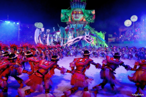 Eine Gruppe von Performern in bunten Kostümen tanzt auf einer Bühne beim Rio-Karneval 2014, mit einer Menge, die auf Stühlen sitzt und festlichen Dekorationen im Hintergrund.