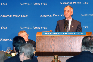 Ein Mann in Blazer, Krawatte und Brille spricht in ein Mikrofon an einem Podium mit einer Tafel, während er einer sitzenden Menge gegenübersteht.
