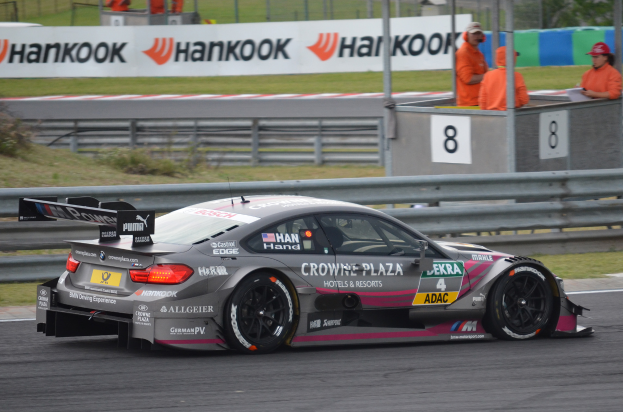 Ein BMW M4 DTM-Rennwagen auf einer Rennstrecke, umgeben von einem Metallzaun, Gras, Pflanzen, einer Gruppe von Menschen, einem Schild mit Text, Pfosten, einem Metallrahmen, einem Zaun, Bäumen und einem bewölkten Himmel.