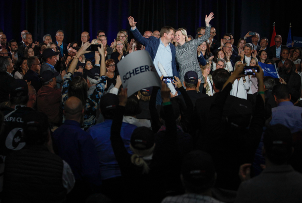 Ein Mann und eine Frau stehen vor einer Menge, einige halten Schilder und Handys, im Hintergrund sind Fahnen und ein Vorhang zu sehen.