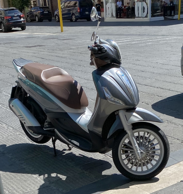 Ein Motorroller mit Helm darauf auf einer Straße geparkt, mit Gebäuden, Bäumen, Pfählen, Menschen, Stühlen, Tischen und anderen Gegenständen im Hintergrund.