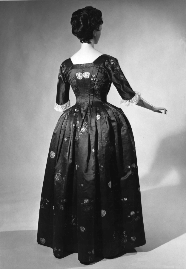 Eine Frau in einem 1950er-Vollständig angepasster Oberteilkleid mit einem langen Rock steht in einem Schwarz-Weiß-Foto, Hände verschränkt, trägt schwarze Pumps und eine klassische Hochsteckfrisur mit dezentem Make-up.