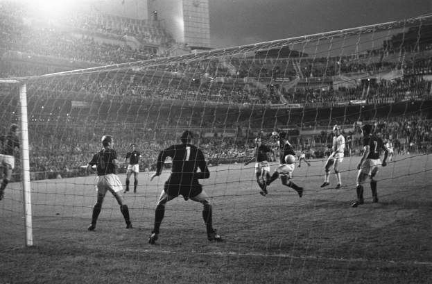 Eine Gruppe von Männern, die Fußball spielen, auf einem Stadionrasen mit einem Tor im Vordergrund und einem Gebäude im Hintergrund, dargestellt in Schwarz-Weiß.