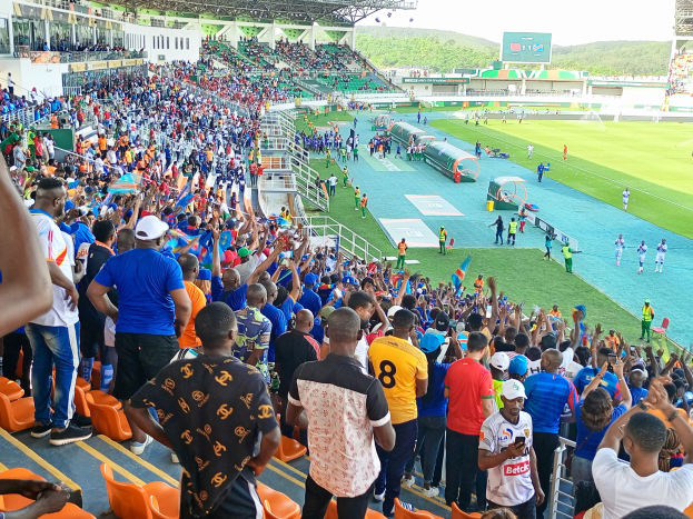 Eine große Menschenmenge versammelt sich vor einem eingezäunten Fußballfeld, wobei einige auf Stühlen sitzen und andere stehen, mit Blick auf Hügel und einen klaren blauen Himmel während der Afrikameisterschaft 2019.