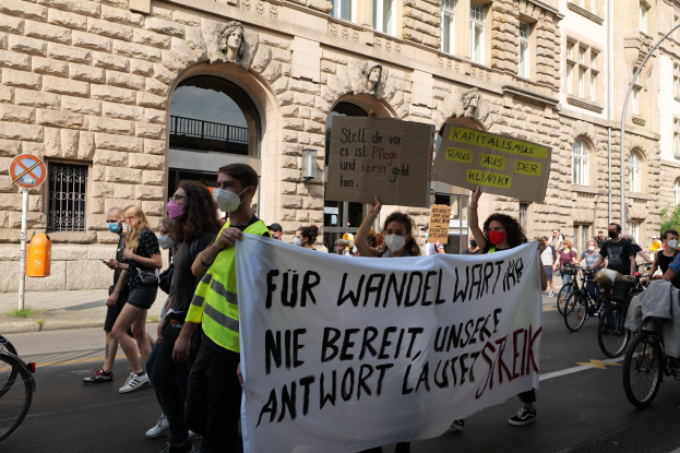 Eine Gruppe von Menschen marschiert bei einer Demonstration gegen die Anti-Kriegsbewegung in Berlin, einige halten Schilder und andere fahren Fahrräder, im Hintergrund ein Gebäude mit Fenstern, Bögen, Säulen und Skulpturen.