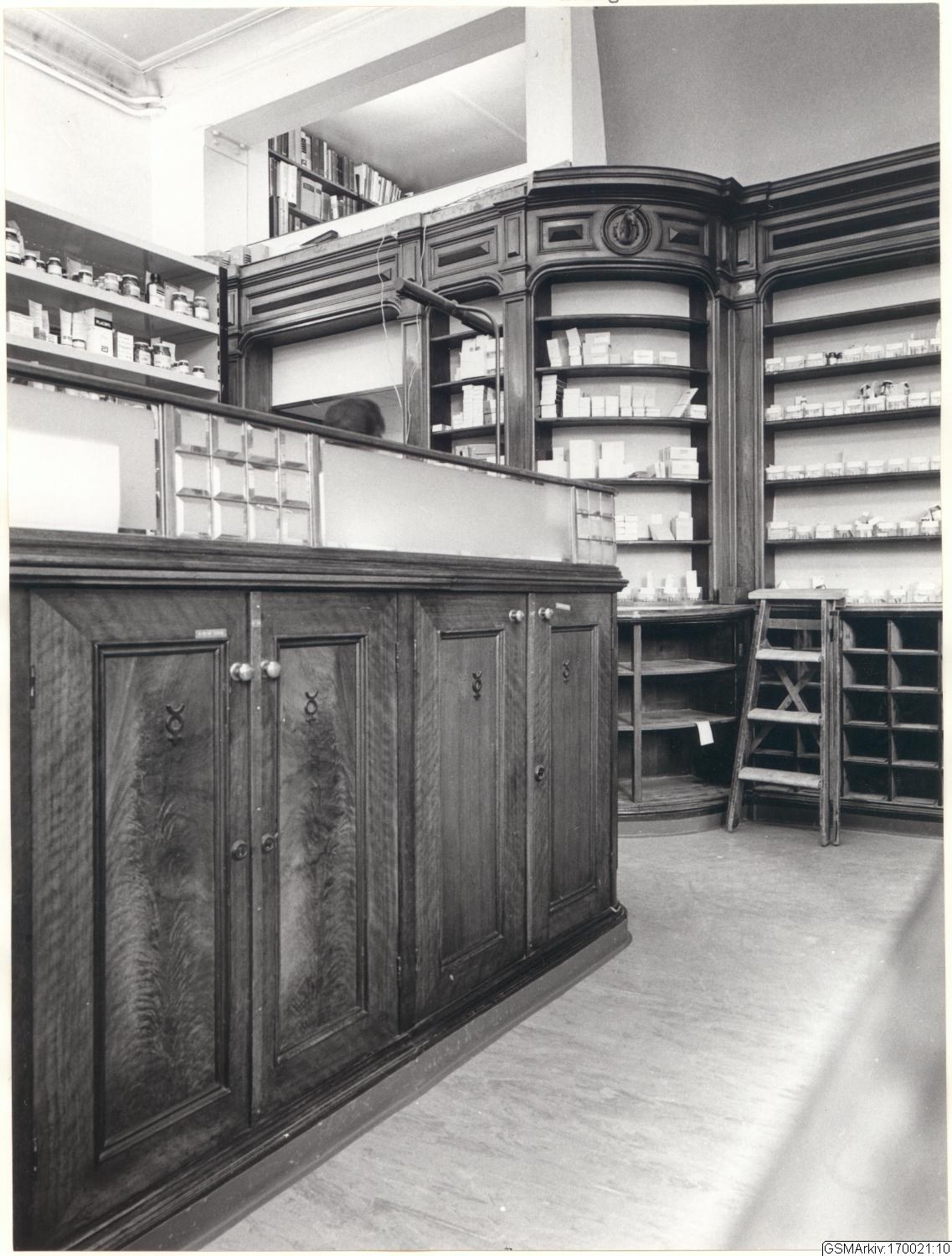 Schwarz-weiß-Foto eines vintage-Apotheken-Interieurs mit einer Theke, Schränken, einer Leiter, Regalen voller Behälter und Text oben.