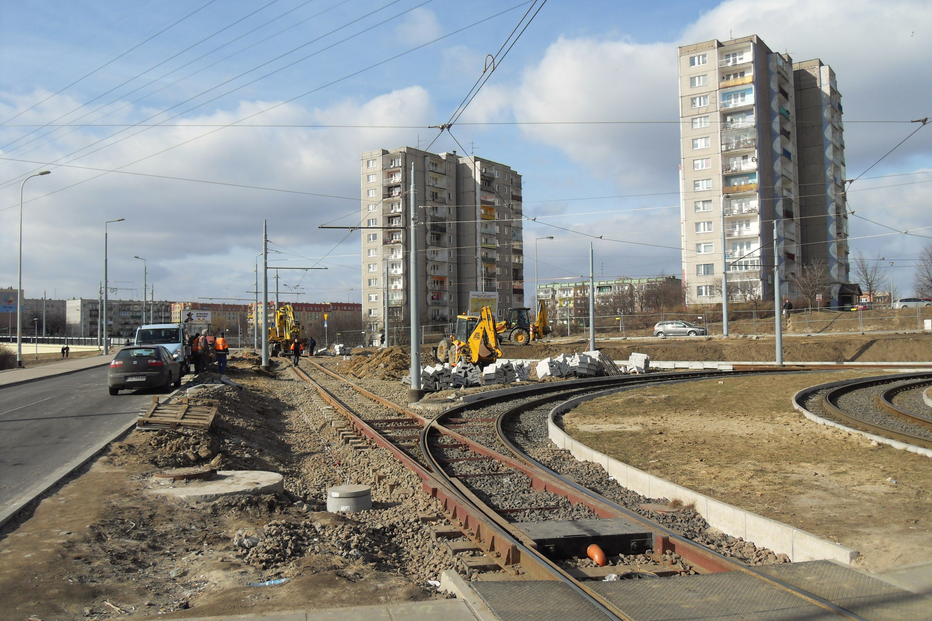 Baustelle mit einer durchlaufenden Bahnschiene, umgeben von Geb├Ąuden, Stra├čeninfrastruktur, Kraftfahrzeugen, Personen, Schutt, B├Ąumen und einem bew├Âlktem Himmel.