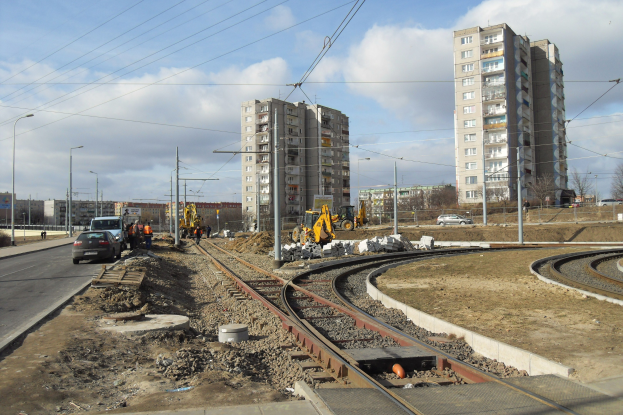 Baustelle mit einer durchlaufenden Bahnschiene, umgeben von Geb├Ąuden, Stra├čeninfrastruktur, Kraftfahrzeugen, Personen, Schutt, B├Ąumen und einem bew├Âlktem Himmel.