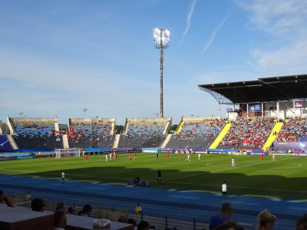 Ein Fußballspiel in einem großen Stadion mit Spielern auf dem Feld und Zuschauern auf den Tribünen unter Flutlicht, mit dem Himmel im Hintergrund.