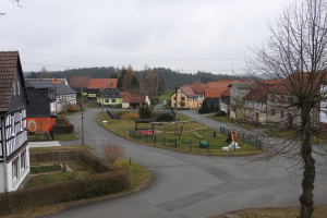 Kleiner Ort mit Häusern, Straßeninfrastruktur, Fahrzeugen, Grünfläche, einem Spielplatz und einem klaren blauen Himmel.