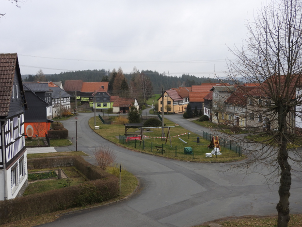 Kleiner Ort mit Häusern, Straßeninfrastruktur, Fahrzeugen, Grünfläche, einem Spielplatz und einem klaren blauen Himmel.