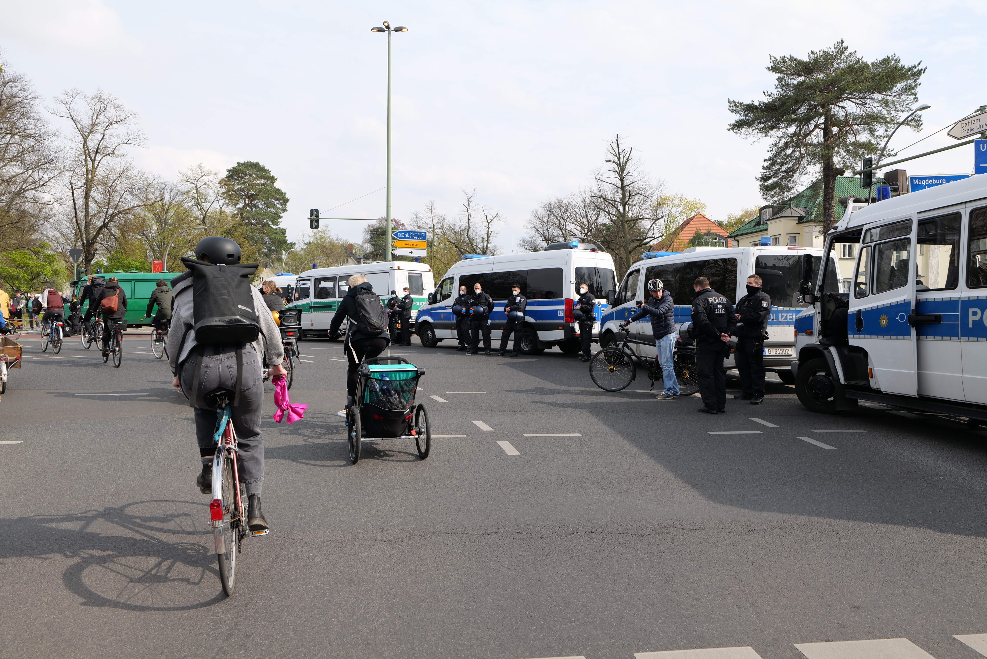 Gruppe von Menschen, die auf Fahrrädern eine Straße entlangfahren, mit Polizeiwagen, einige tragen Helme und haben Taschen, andere schieben Kinderwagen, vor einem Hintergrund aus Bäumen, Gebäuden und Verkehrsinfrastruktur.