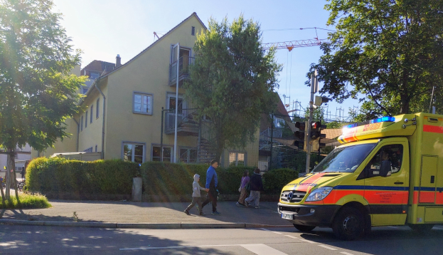 Ambulanz am Straßenrand geparkt mit Menschen auf dem Gehweg, Ampeln, Pfosten, Bäume, Pflanzen, Gras, Gebäude mit Fenstern und einen Kran im Hintergrund unter einem klaren blauen Himmel.