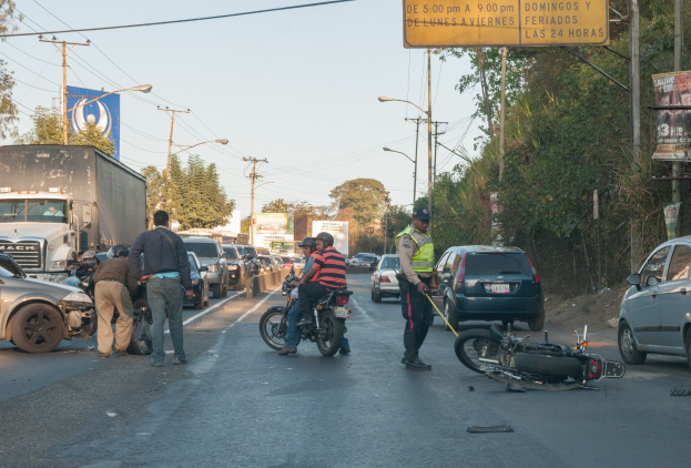 Gruppe von Menschen um ein verunglücktes Motorrad auf der Fahrbahn mit mehreren Fahrzeugen, darunter ein Lastwagen, und Hintergrund-Elementen wie Bäumen, Pfählen, Lampen, Schildern und Himmel.