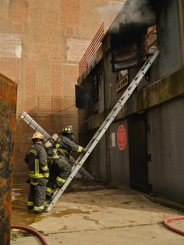 Feuerwehrleute in Helmen klettern an einer Leiter zu einem rauchgefüllten Gebäude, mit einem Metallobjekt links und Rohren am Boden.