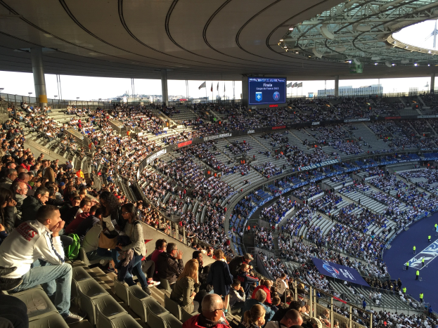 Große Menschenmenge in einem Stadion bei einem Fußballspiel, mit einer Bühne, Fahnen, Stangen, einem Bildschirm und der Allianz Arena in München im Hintergrund.