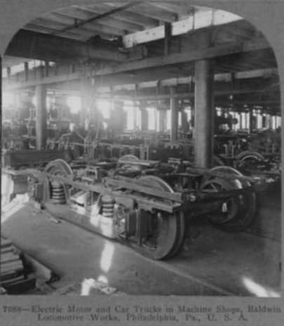 Schwarzes und weißes Foto eines Elektromotors und eines Lastwagens in einer Fabrik mit Maschinen, Säulen und einem Dach, mit Text unten.