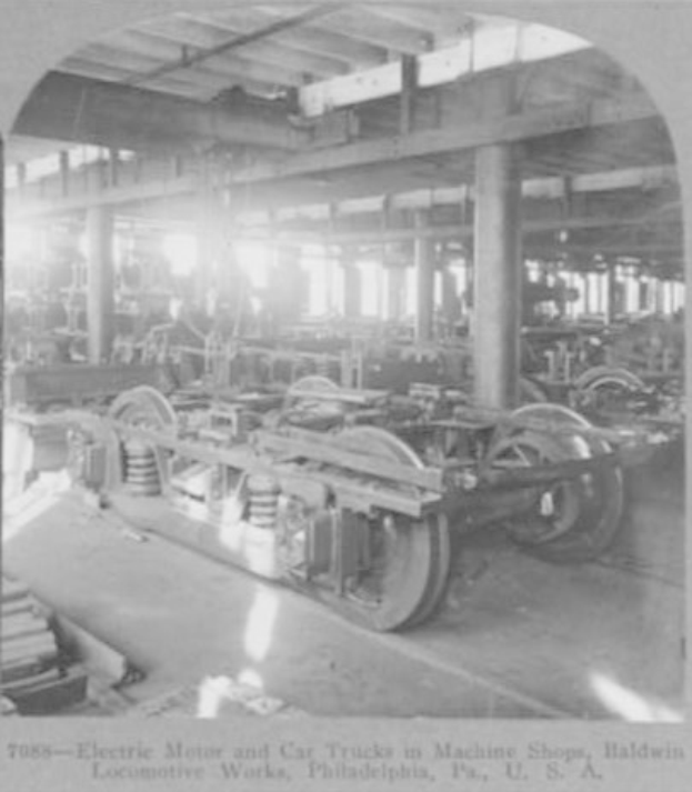 Schwarzes und weißes Foto eines Elektromotors und eines Lastwagens in einer Fabrik mit Maschinen, Säulen und einem Dach, mit Text unten.