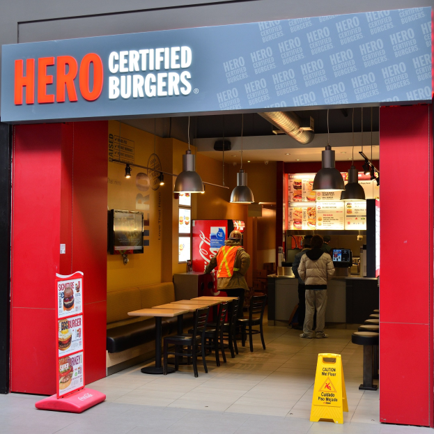 Außenansicht eines Hero Certified Burgers Restaurants in London mit Menschen drinnen, Tischen, Stühlen, einer Speisekarte, einem wandmontierten Fernseher und Deckenleuchten.