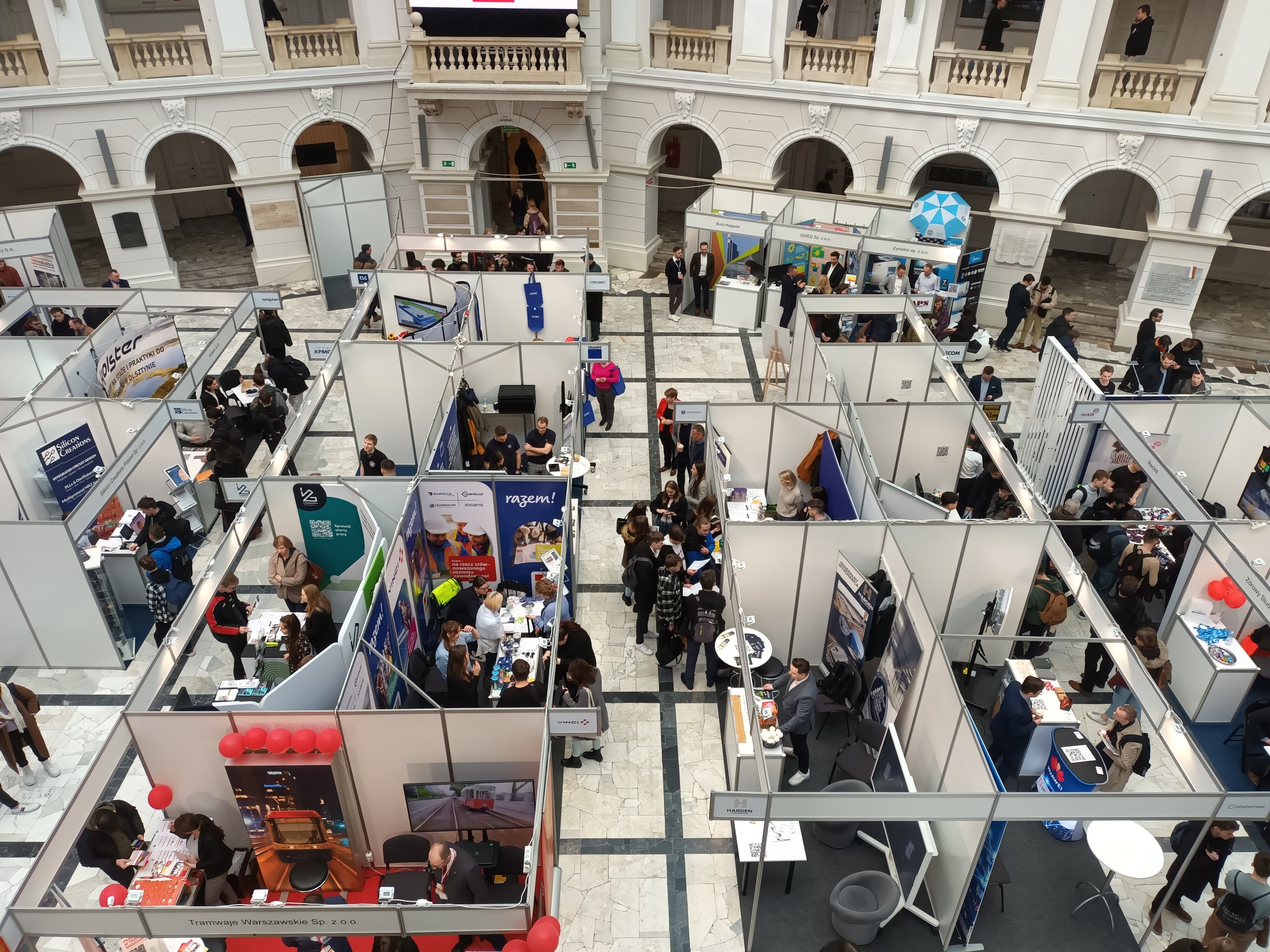 Eine große Gruppe von Menschen auf einer internationalen Fachmesse mit Ständen, Bannern, Tischen, Stühlen und Ballons, vor einem Gebäude mit Bögen, Säulen, Geländern und einem Bildschirm.