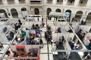 Eine große Gruppe von Menschen auf einer internationalen Fachmesse mit Ständen, Bannern, Tischen, Stühlen und Ballons, vor einem Gebäude mit Bögen, Säulen, Geländern und einem Bildschirm.