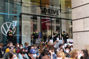 Große Gruppe von Menschen mit Masken und Schildern vor einem Gebäude mit Glaswänden und Text, einige tragen Mützen und halten Kameras während einer Demonstration in Melbourne, Australien.