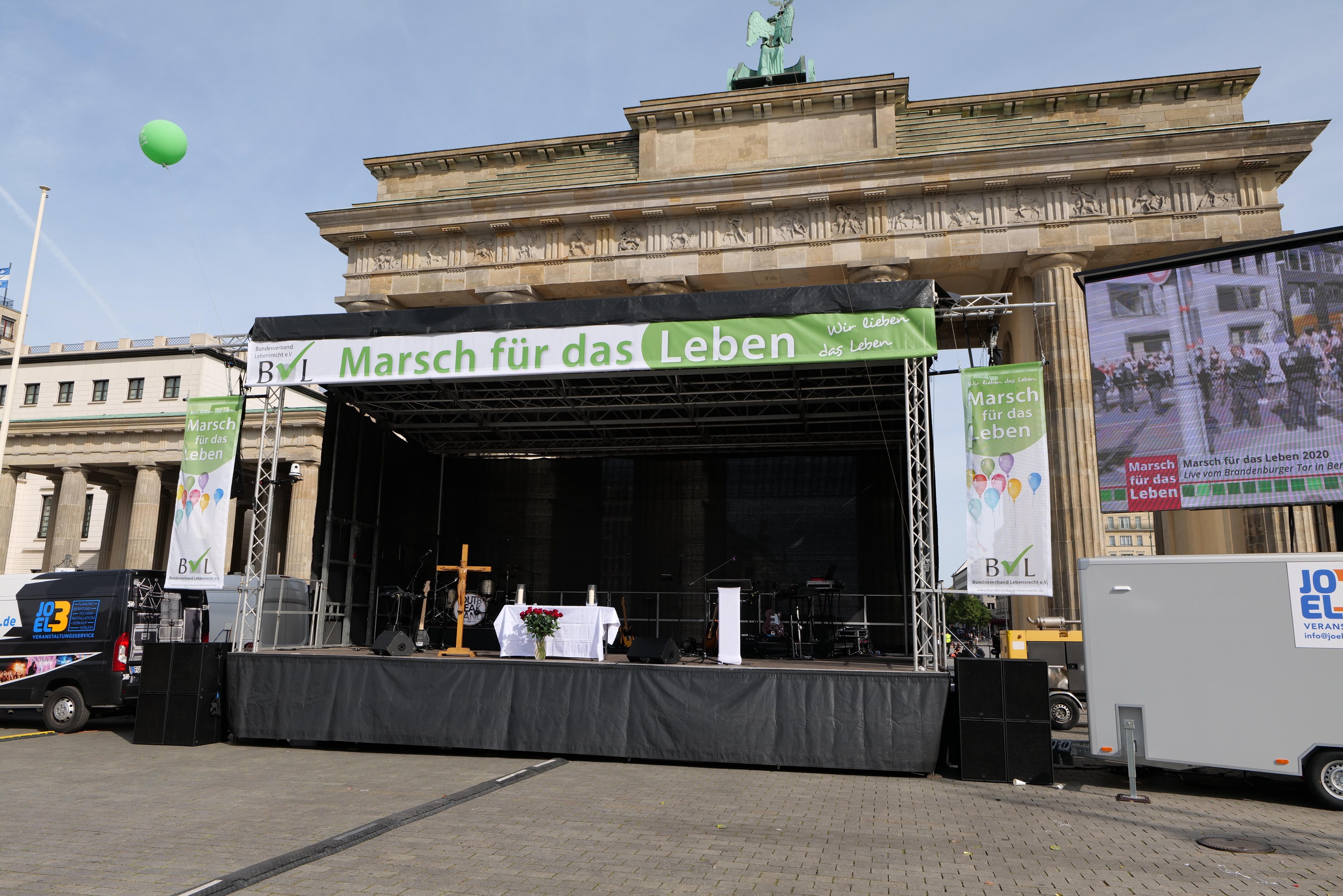 Bühne vor dem Brandenburger Tor in Berlin, Deutschland, mit einem Tisch mit einer Blumenvase, einem Kreuzsymbol, Transparenten mit Text, Lautsprechern und anderen Gegenständen, sowie Fahrzeugen auf der Straße und Gebäuden im Hintergrund.