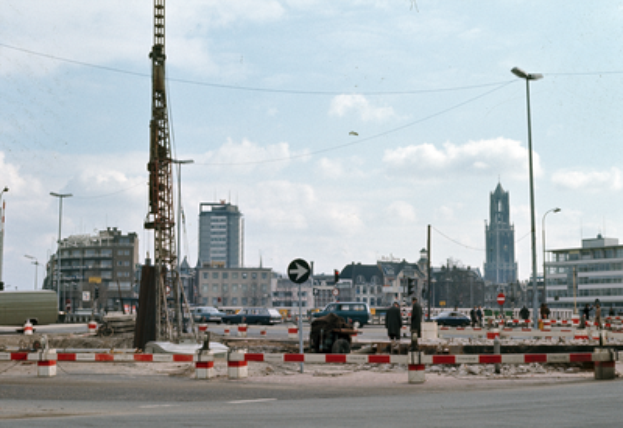 Eine Baustelle mit einem großen zentralen Turm, umgeben von Gebäuden, Straßenmasten, Straßenlaternen, Kraftfahrzeugen, Absperrpfeilern, Schildern, Strommasten, Stromkabeln und einer wolkigen Himmel.