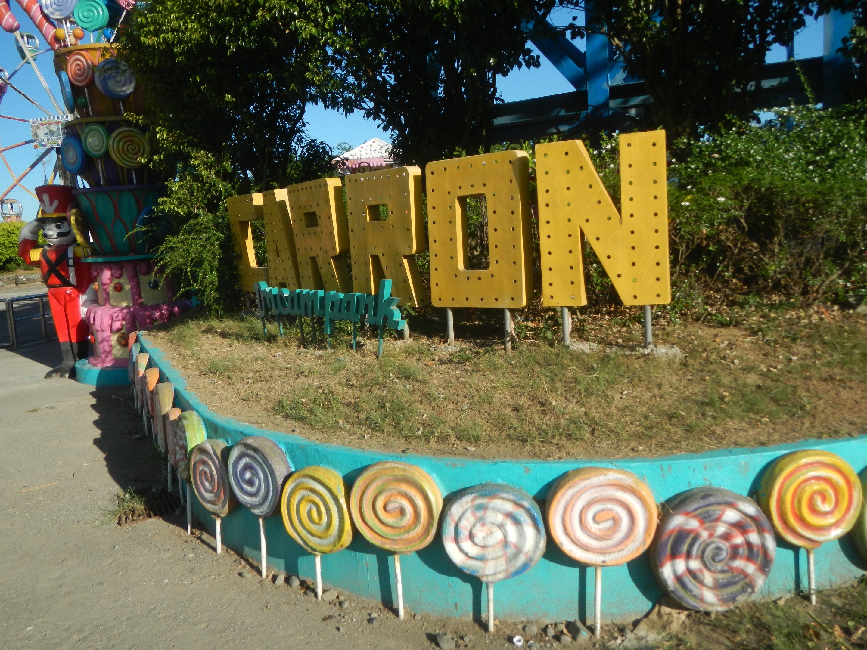Karnevalszenen mit einem großen Riesenrad im Hintergrund, Bäumen, Gras, einer Person links, einer Straße unten, Lollis und einem Namensschild im Vordergrund und Himmel oben.
