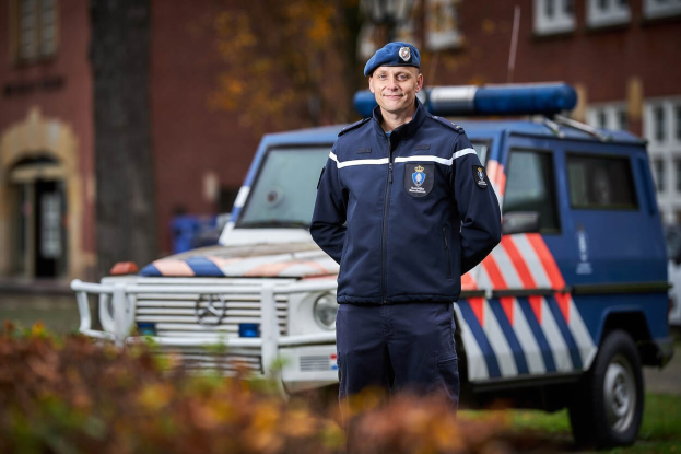 Polizeibeamter in Uniform vor einem Polizeiwagen mit leicht unscharfem Hintergrund und Pflanzen unten.