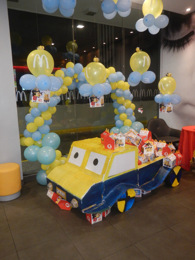 Eine McDonald's Geburtstagsfeier mit einem gelben und blauen Lastwagen, der mit Ballons und anderen Dekoartikeln geschmückt ist, einem rot eingedeckten Tisch mit Gegenständen, Hockern und einer Glaswand im Hintergrund, sowie einer Decke mit Lichtern, die eine festliche Atmosphäre schaffen.