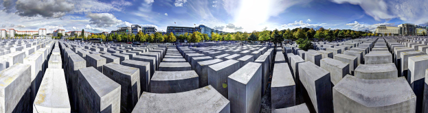 Luftaufnahme des Holocaust-Mahnmals für die ermordeten Juden Europas in Berlin, mit großen Betonplatten, Bäumen, umgeben von Gebäuden, Fahrzeugen auf einer Straße und einem bewölkten Himmel.