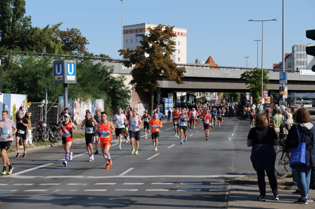 Gruppe von Menschen, die bei einem Marathon auf einer baumbestandenen Straße laufen, mit Laternenpfählen, Schildern, Fahrrädern, einem Zaun, Gras, einer Brücke, Gebäuden und einem klaren blauen Himmel.