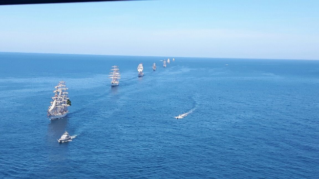 Eine große Flotte von Segelschiffen segelt über einen hellblauen Ozean unter einem klaren Himmel und nimmt an einem Regatta teil.