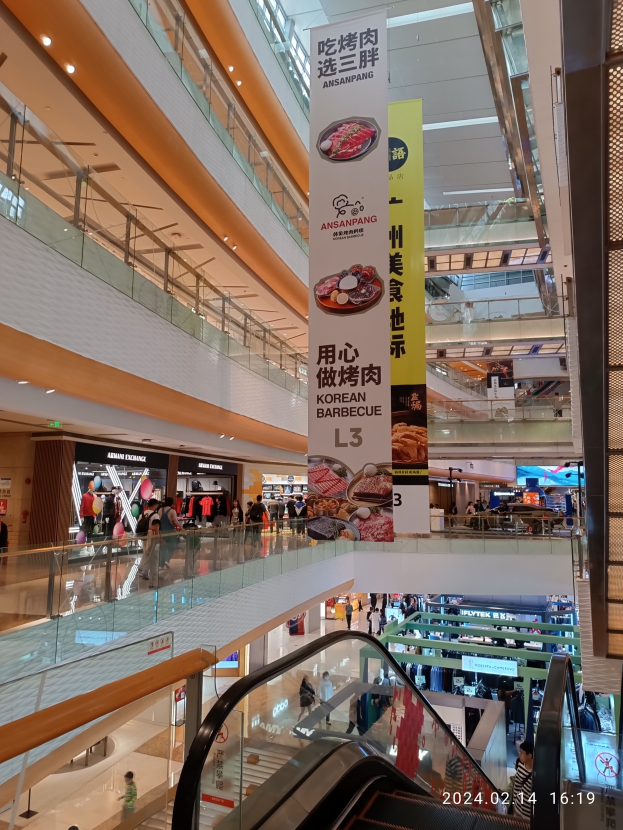 Einblicke in ein Einkaufszentrum mit einer Rolltreppe in der Mitte, Menschen, die herumlaufen, einige mit Handtaschen, und Schilder mit Text an der Decke, beleuchtet von Lichtern mit Geschäften im Hintergrund.