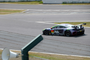 Ein Rennsportwagen fährt auf einer Rennstrecke mit einer Absperrung im Hintergrund, umgeben von Gras und einem Lautsprecher unten, mit einem unscharfen Hintergrund, der Bewegung suggeriert.