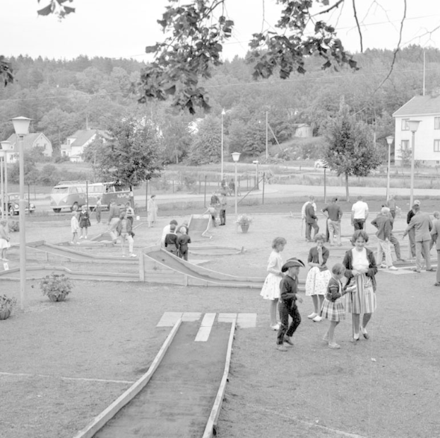 Schwarz-weißes Bild von Menschen, die Minigolf in einem Park mit üppiger Vegetation, Laternenmasten, geparkten Fahrzeugen und Gebäuden im Hintergrund unter einem sichtbaren Himmel spielen.