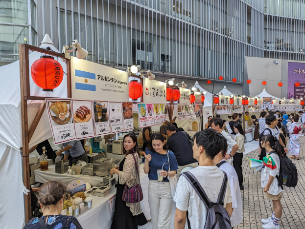 Eine Gruppe von Menschen auf einem Food-Festival in Japan, die um einen Tisch mit Essen versammelt sind, einige tragen Taschen und halten Gläser, mit Schildern, Lichtern und Laternen an der Decke hängend und ein Gebäude im Hintergrund sichtbar.