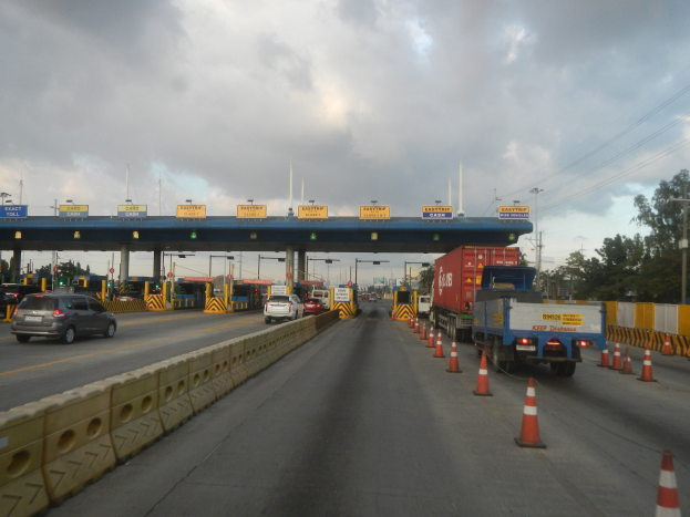 Mautüberwachungsstation mit fahrenden Fahrzeugen, Absperrgittern auf der Stra├če, eine Trennwand, ein Tor mit Namensschildern, Laternenpf├Ąhle, Strommasten mit Kabeln, B├Ąume und ein bew├Âlktes Himmel.
