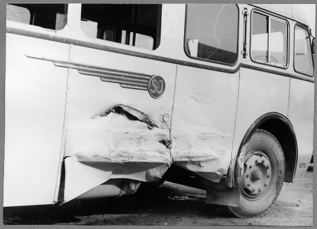 Schwarz-weiß-Foto eines beschädigten Busses am Straßenrand mit eingedellter Front, zerbrochenen Fenstern und offener Fahrertür, die das Innere zeigt.