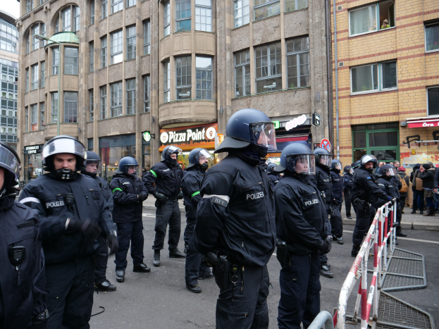 Eine Gruppe von Polizeibeamten vor einer Menge hinter Absperrungen auf einer Straße stehend, mit Gebäuden, Texttafeln, Laternenpfählen und einem klaren blauen Himmel im Hintergrund.