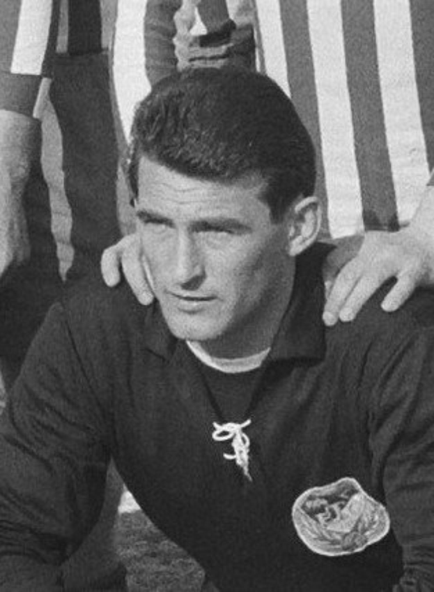 Schwarzes und weißes Foto eines Mannes, der auf einem Rasenfussballfeld sitzt und ein T-Shirt und eine Kette trägt, mit zwei Personen dahinter.