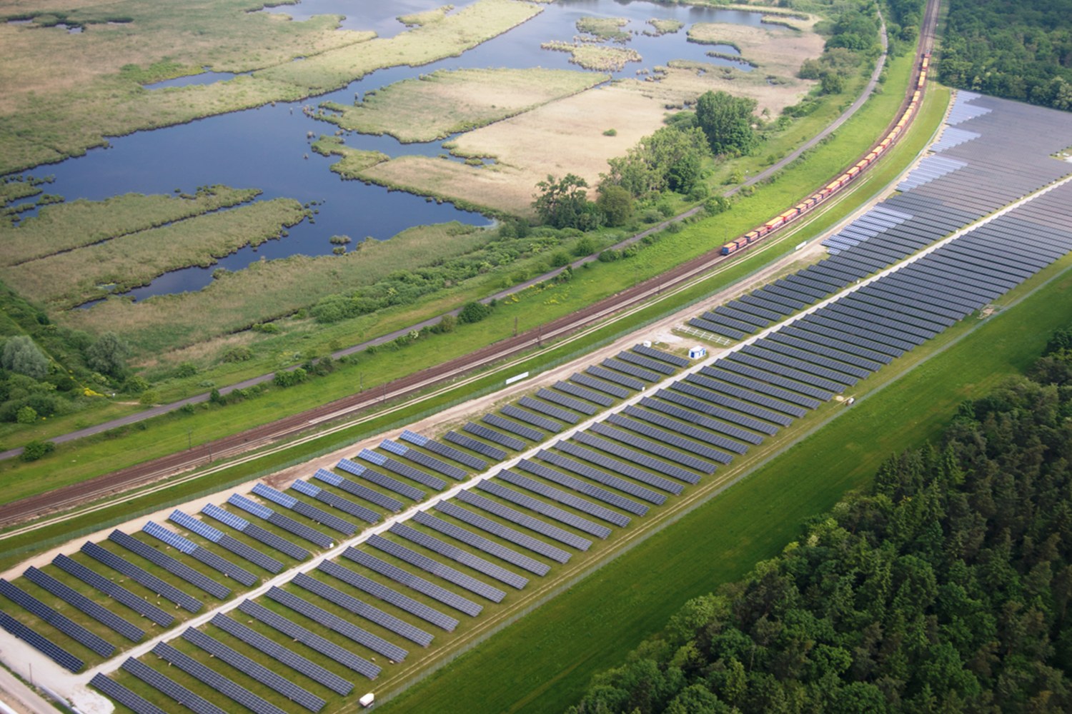 Luftaufnahme einer Solaränlage mit Panelen, umgeben von Bäumen, Gras, Wasser und einer nahen Bahntrasse.