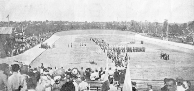Schwarzes und weißes Foto von Zuschauern bei einem Pferderennen in einem Stadion, einige sitzen auf Bänken, andere stehen oder reiten Pferde, mit Bäumen und wolkenlosem Himmel im Hintergrund.