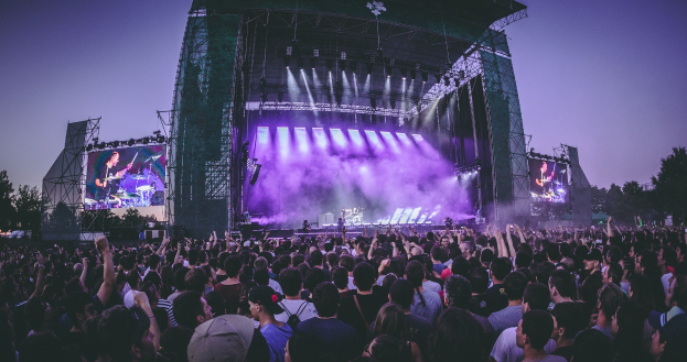 Eine große Menge bei einem Musikfestival schaut auf einer hell erleuchteten Bühne mit Instrumenten und Bildschirmen, die vor einem Gebäude, Bäumen und einem klaren blauen Himmel aufgebaut sind.