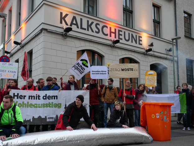 Gruppe von Menschen mit Protestschildern und Plakaten vor einem Gebäude, mit zwei Personen im Vordergrund und einem Müllcontainer rechts.