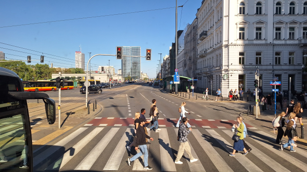 Gruppe von Menschen, die eine Straße an einer Ampel überqueren, mit einem Bus auf der linken Seite, Fahrzeugen auf der Straße, Verkehrsampeln, Laternenmasten, Gebäuden, Bäumen und einem klaren blauen Himmel.