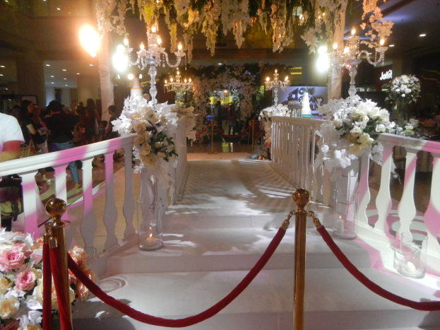 Eine Hochzeitszeremonie in einem Einkaufszentrum mit weißem Teppich, dekorativen Geländern, Blumenarrangements und Deckenbeleuchtung, bei der eine Menschenmenge anwesend ist, darunter auch Personen mit Taschen.