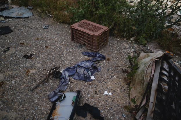 Haufen Müll, einschließlich einer Plastikbox und Kleidung neben einem Zaun, mit Vegetation im Hintergrund.