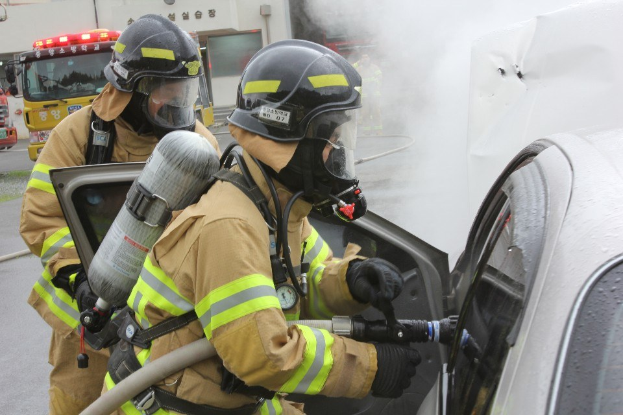 Zwei Feuerwehrleute in Schutzausröstung verwenden einen Schlauch, um ein brennendes Auto zu löschen, mit Rauch, der aus dem Fahrzeug aufsteigt und andere Fahrzeuge und ein Gebäude im Hintergrund zu sehen sind.
